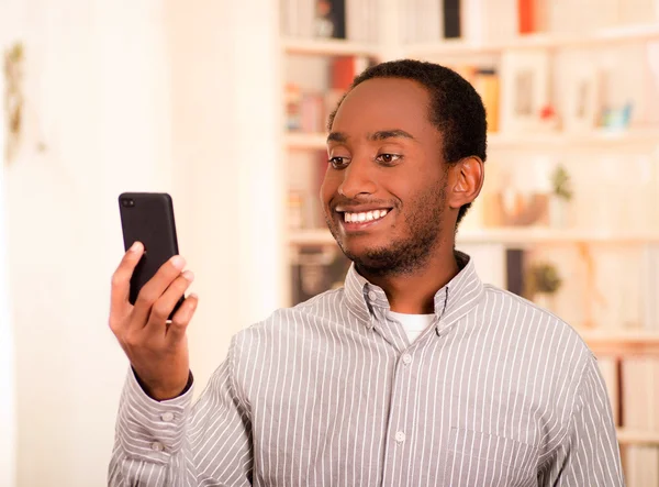 Handsome man wearing casual clothes holding up mobile phone looking at screen and smiling