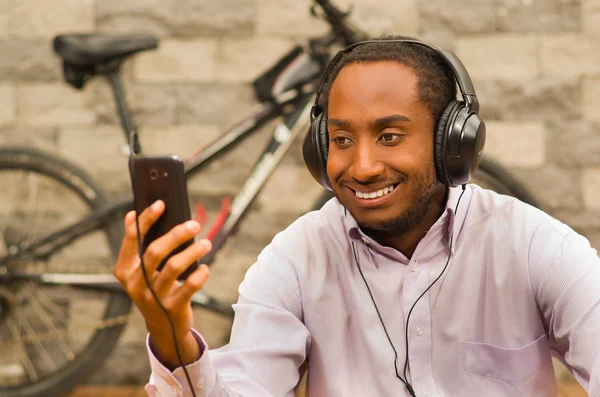 Man wearing white red business shirt sitting down with headphones on, enjoying music while looking at mobile screen, smiling happily, bicycle standing behind leaning against grey brick wall