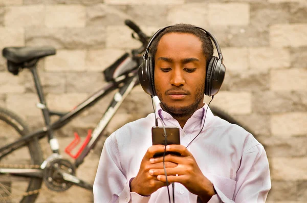 Man wearing white red business shirt sitting down with headphones on, enjoying music while looking at mobile screen, bicycle standing behind leaning against grey brick wall