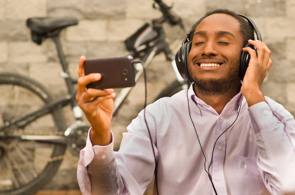 Man wearing white red business shirt sitting down with headphones on, enjoying music while looking at mobile screen, smiling happily, bicycle standing behind leaning against grey brick wall
