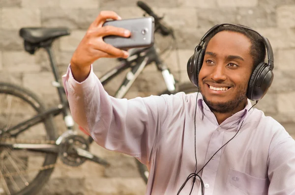 Man wearing white red business shirt sitting down, headphones on head, holding up mobile phone taking selfie photo, smiling and posing, bicycle standing behind leaning against grey brick wall