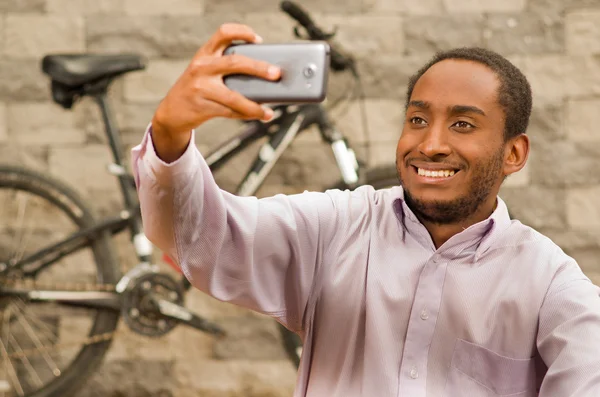 Man wearing white red business shirt sitting down, holding up mobile phone taking selfie photo, smiling and posing, bicycle standing behind leaning against grey brick wall