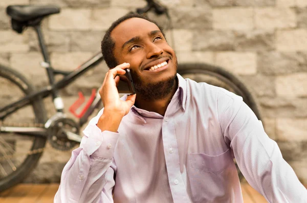 Man wearing white red business shirt sitting down, smiling and talking on mobile phone, bicycle standing behind leaning against grey brick wall