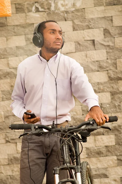 Man wearing grey office pants, white red business shirt standing by bicycle holding mobile phone, headphones on head, smiling and posing, brick wall background