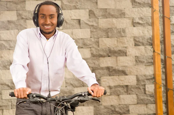 Man wearing grey office pants, white red business shirt standing by bicycle holding mobile phone, headphones on head, smiling and posing, brick wall background