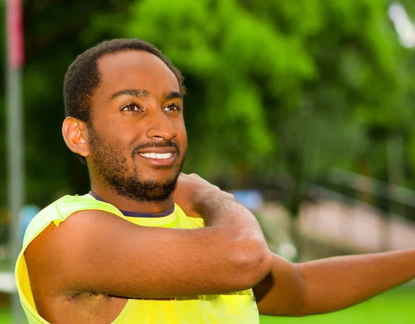 Man wearing yellow shirt stretching arms in park sorrounded by green grass and trees, training concept