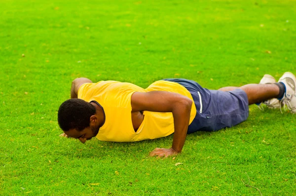 Man wearing yellow shirt and blue shorts doing push ups using arms in park sorrounded by green grass trees, training concept