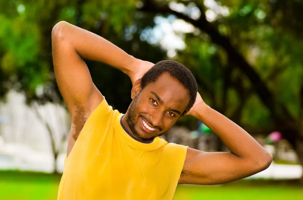 Man wearing yellow shirt stretching arms behind head in park sorrounded by green grass and trees, training concept