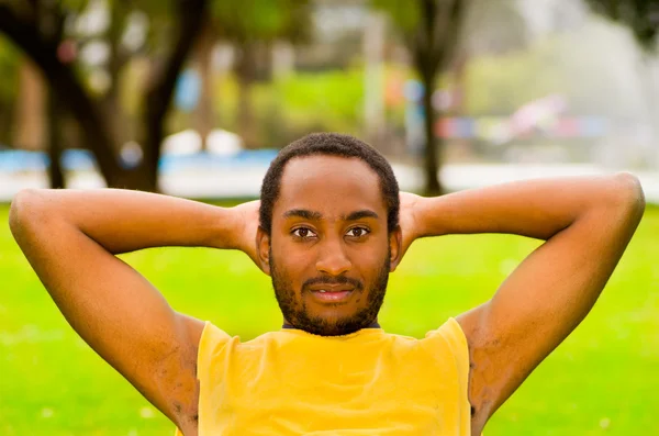 Man wearing yellow shirt doing situps in park sorrounded by green grass and trees, happy facial expression, training concept