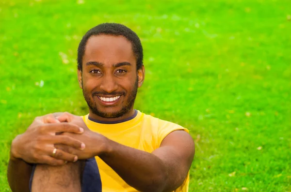 Man wearing yellow shirt and blue shorts sitting on green grass, holding arm around knee smiling to camera, training concept