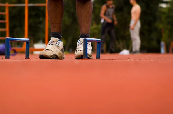 White shoes with mans legs standing on orange athletic running surface between two florr mounted blue handles, blurry people training background