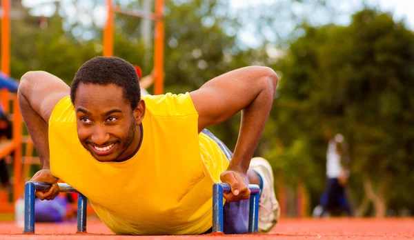 Man wearing yellow shirt doing push-ups at outdoors training facility, orange athletic surface and green trees background