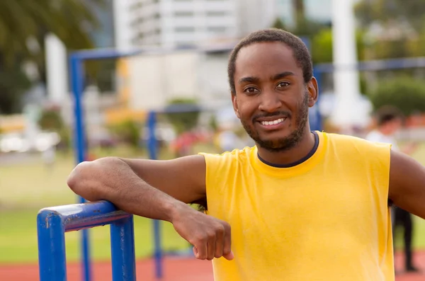 Man wearing yellow shirt and blue shorts smiling while posing for camera at outdoors training facility with orange athletic surface, blurry people background