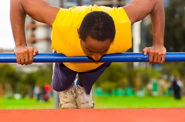Man wearing yellow shirt and blue shorts doing static strength excercises hanging from pole, outdoors training facility with orange athletic surface, blurry people background