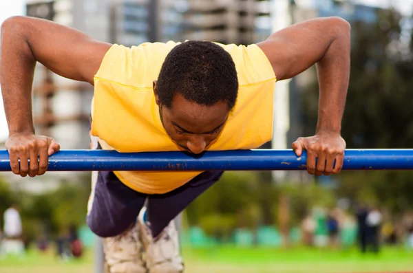 Man wearing yellow shirt and blue shorts doing static strength excercises hanging from pole, outdoors training facility with orange athletic surface, blurry people background