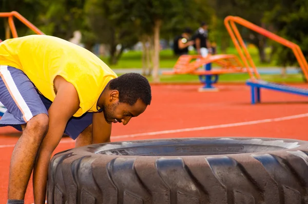 Man wearing yellow shirt and blue shorts lifting large tractor tire during strength excercise, outdoors training facility with orange athletic surface, green trees background