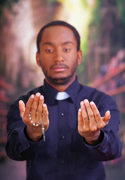 Catholic priest wearing traditional clerical collar shirt standing facing camera, holding hands out with rosary cross, looking forward, religion concept