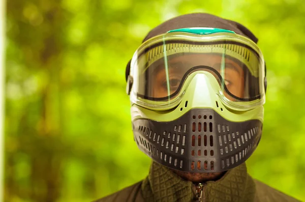 Closeup headshot man wearing jacket, green and black protection facial mask standing facing camera, forest background, paintball concept