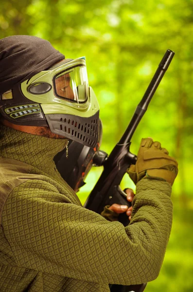 Closeup headshot man wearing jacket, green and black protection facial mask standing in profile angle with weapon, forest background, paintball concept