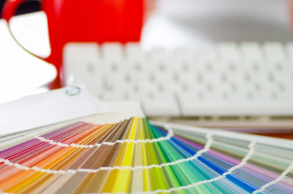 Closeup large palette colormap spread out in front of white computer keyboard and red coffee mug sitting on wooden desk, designer concept