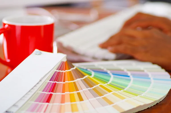 Closeup large palette colormap spread out in front of white computer keyboard and red coffee mug sitting on wooden desk, designer concept