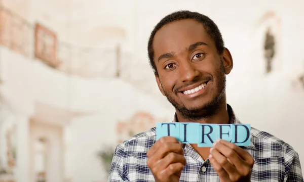 Headshot handsome man holding up small letters spelling the word tired and smiling to camera
