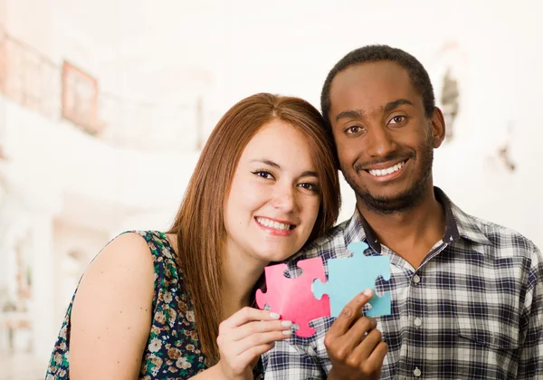 Interracial charming couple embracing friendly, holding up large puzzle pieces and happily interacting having fun, blurry studio background
