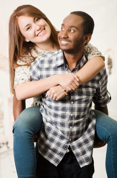 Interracial couple wearing casual clothes interacting having fun, man carrying woman on his back, both smiling happily, white studio background