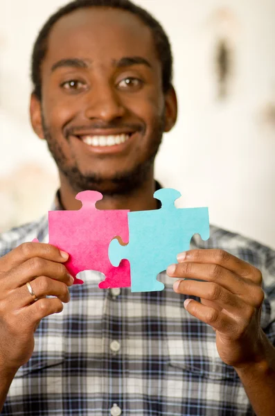 Headshot handsome man holding up big puzzle pieces in pink and blue, smiling happily to camera, white studio background