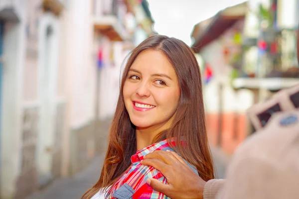 Pretty brunette woman turns around smiling while boyrfriends hand touches her shoulder, vibrant colors and blurry urban background