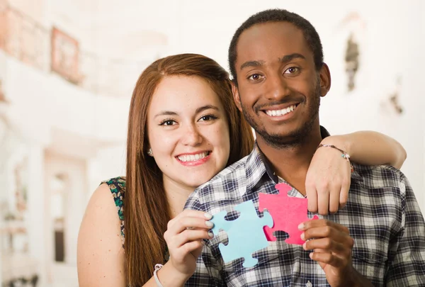 Interracial charming couple embracing friendly, holding up large puzzle pieces and happily interacting having fun, blurry studio background