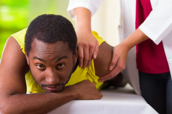 Man lying down getting physical shoulder treatment from physio therapist, patient looking into camera while her hands working on his upper arm area, medical concept