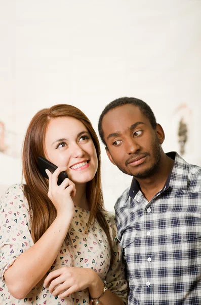 Interracial charming couple wearing casual clothes posing interacting friendly, woman talking on cell phone and man listening in, white studio background