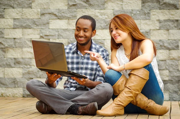 Interracial charming couple wearing casual clothes sitting down on wooden surface lookin at laptop together, in front of grey brick wall