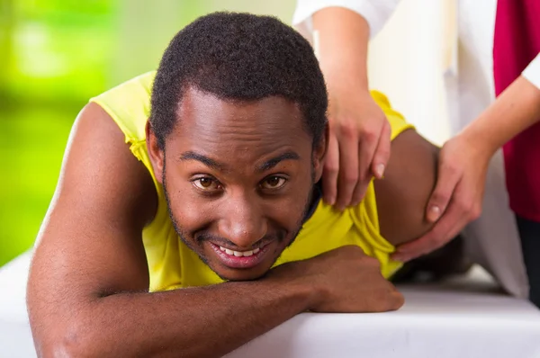 Man lying down getting physical shoulder treatment from physio therapist, patient looking into camera while her hands working on his upper arm area, medical concept