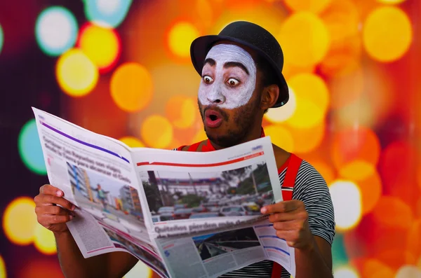 Pantomime man with facial paint posing for camera holding newspaper showing funny face, blurry lights background