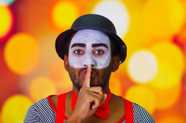 Headshot pantomime man with facial paint posing for camera using hands to cover mouth, blurry lights background