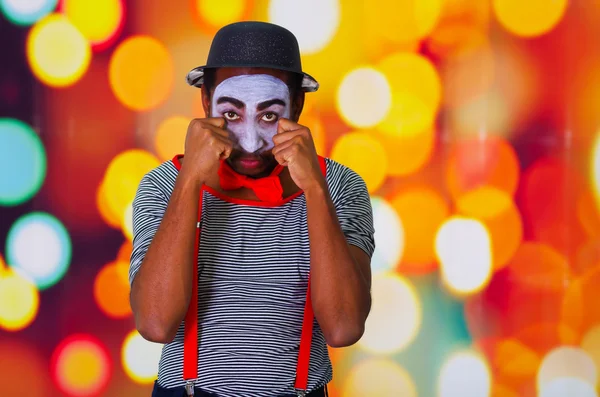 Headshot pantomime man with facial paint posing for camera using hands interacting rubbing eyes, blurry lights background