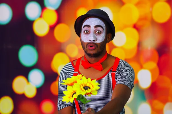 Headshot pantomime man with facial paint posing for camera holding sunflowers in hands, blurry lights background