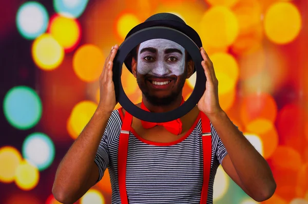 Pantomime man with facial paint posing for camera holding circle showing face in middle, blurry lights background