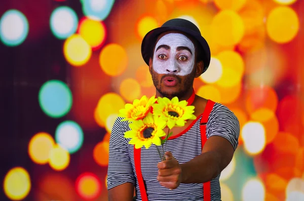 Headshot pantomime man with facial paint posing for camera holding sunflowers in hands, blurry lights background