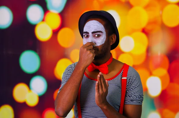 Pantomime man wearing facial paint posing for camera, using hands interacting body language, blurry lights background