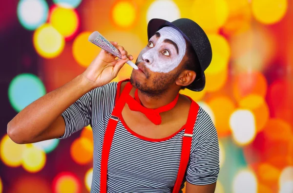 Headshot pantomime man with facial paint posing for camera using blow horn, blurry lights background