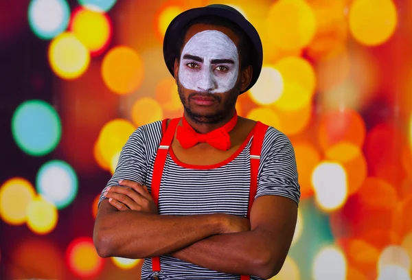 Pantomime man wearing facial paint posing for camera, standing with arms crossed, blurry lights background