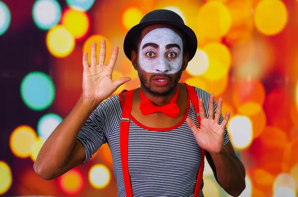 Pantomime man with facial paint posing for camera interacting funny using hands, blurry lights background