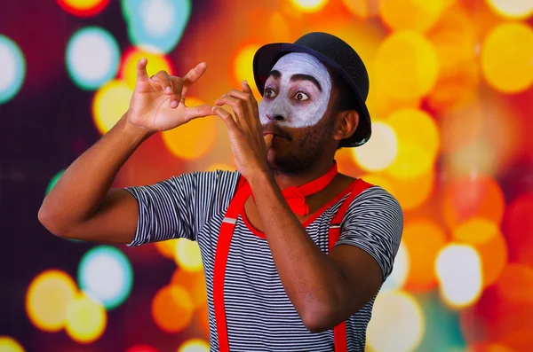 Pantomime man wearing facial paint posing for camera, using hands interacting body language, blurry lights background