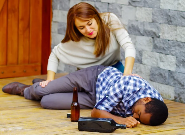 Man wearing casual clothes lying drunk passed out on wooden surface, pretty woman sitting beside him trying to get contact by touching and shaking