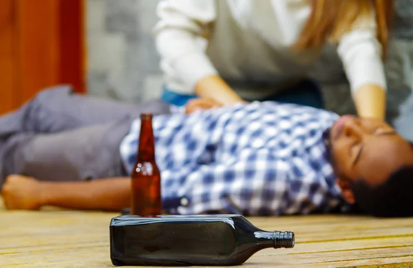 Man wearing casual clothes lying drunk passed out on wooden surface, pretty woman sitting beside him trying to get contact by touching and shaking