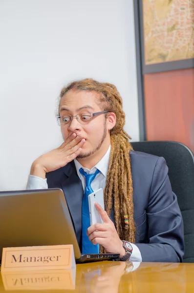 Handsome man with dreads and business suit sitting by desk, holding mobile phone while looking at laptop, young manager concept