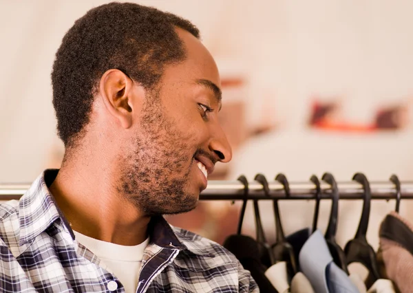 Headshot young man smiling to camera, standing in front of clothing rack, fashion concept
